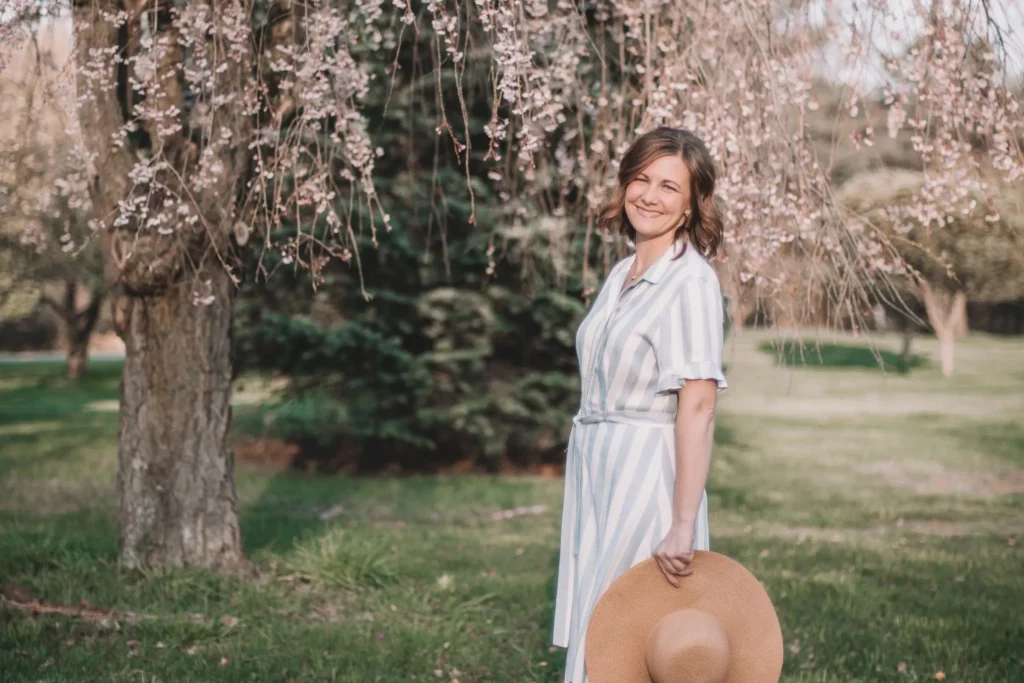 Molly Ijames smiling under blossoming tree branches, wearing a light blue and white striped dress and holding a wide-brimmed straw hat in a sunlit park.