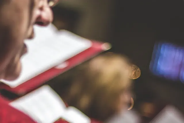Close-up of choir members singing from sheet music during a rehearsal or performance, with a soft focus on the background and warm, ambient lighting.
